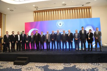 First image shows a man in a suit speaking at a podium with a microphone in front of a blue backdrop featuring the Gün Sazak name and themes of merit and honesty in public service, alongside a large portrait of Gün Sazak wearing glasses and a mustache. Second image depicts a man in a suit holding a microphone at a podium with a wooden backdrop and a large black-and-white portrait of Gün Sazak, text reading Gün Sazak Makale Yarışması and Kamuda Liyakat ve Dürüstlük. Third image displays a man in a suit speaking at a podium with a microphone, blue backdrop with Türk Büro-Sen logo and Gün Sazak portrait, text on merit and honesty in public service. Fourth image features a group of about 15 people in formal suits and dresses standing on a stage with a blue backdrop displaying a circular emblem.
