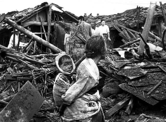 Black-and-white close-up photo of a distressed Korean woman carrying a young child on her back amid wooden debris and collapsed structures; the child wears a hooded garment, and the scene shows emotional hardship in a destroyed residential area.