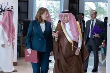 First image shows a man in traditional Saudi attire shaking hands with a woman in a blue suit against Saudi and UK flags in a formal setting with shelves and decor. Second image depicts a long conference table with multiple attendees in business and traditional clothing seated around laptops documents and refreshments under Saudi and UK flags in a modern room. Third image captures the woman in blue suit and man in Saudi attire walking and gesturing in discussion with other suited men in background holding folders near flags and decor.