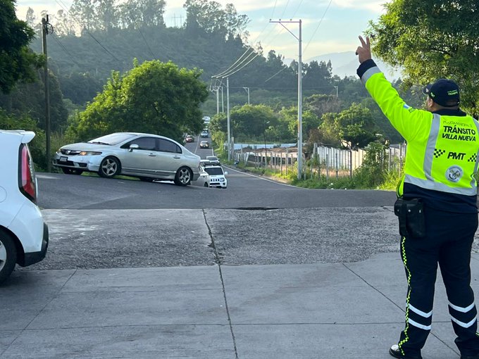 Traffic officer in yellow high-visibility vest and pants with PMT patches stands on asphalt road directing with raised right arm and pointing finger wearing black cap and utility belt beside white patrol vehicle gray sedan and small white car positioned at intersection surrounded by green trees utility poles and distant hills under partly cloudy sky