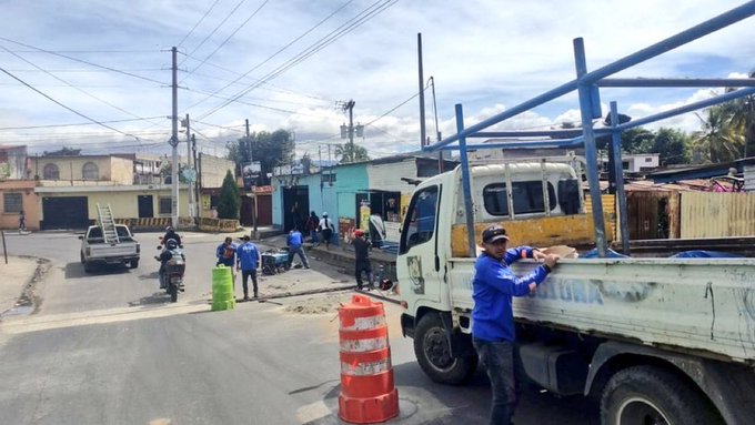 Street scene in a residential area with colorful houses, utility poles, and palm trees in the background. Several workers in blue uniforms stand near a white truck loaded with materials and covered items. Orange traffic cones and barriers are placed around the work zone on the asphalt road. A motorcycle and cars are parked nearby, with one worker wearing sunglasses and a hat adjusting items on the truck.
