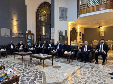 First image shows two men in suits sitting in armchairs facing each other in a room with white walls, a portrait of Mustafa Kemal Atatürk on the wall, and flags of Iraq, Israel, Turkey, and Turkmeneli on stands beside them, with a small table and decorative lights. Second image depicts a group of about eight men in suits seated around wooden tables with tea sets in a large arched room with traditional decor, portraits on walls including Atatürk, and ornate furniture. Third image features two men in suits holding a framed artwork depicting a blue-domed building surrounded by people, standing in front of the Atatürk portrait with the same flags visible. Fourth image shows three men in suits, one handing a red box with a crescent emblem to another, with the Atatürk portrait, Turkish flag, and a blue flag with stars in the background.