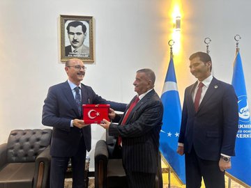 First image shows two men in suits sitting in armchairs facing each other in a room with white walls, a portrait of Mustafa Kemal Atatürk on the wall, and flags of Iraq, Israel, Turkey, and Turkmeneli on stands beside them, with a small table and decorative lights. Second image depicts a group of about eight men in suits seated around wooden tables with tea sets in a large arched room with traditional decor, portraits on walls including Atatürk, and ornate furniture. Third image features two men in suits holding a framed artwork depicting a blue-domed building surrounded by people, standing in front of the Atatürk portrait with the same flags visible. Fourth image shows three men in suits, one handing a red box with a crescent emblem to another, with the Atatürk portrait, Turkish flag, and a blue flag with stars in the background.