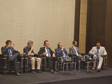 First image displays six men in suits and lanyards seated on a stage with microphones in a conference room featuring beige walls and modern chairs. Second image shows a projector screen with Turkish text about education for Syrian students a male speaker at podium and audience members including women in headscarves seated around tables in a hotel ballroom with Marriott branding. Third image captures a workshop session with diverse attendees in business attire at round tables with laptops documents and water bottles under a chandelier in a spacious room with multiple screens displaying text. Fourth image depicts another meeting with participants seated at tables facing dual screens showing Arabic text a man speaking at podium and photographers capturing the scene in a elegant venue with beige decor and beverages on tables.