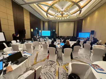 First image displays six men in suits and lanyards seated on a stage with microphones in a conference room featuring beige walls and modern chairs. Second image shows a projector screen with Turkish text about education for Syrian students a male speaker at podium and audience members including women in headscarves seated around tables in a hotel ballroom with Marriott branding. Third image captures a workshop session with diverse attendees in business attire at round tables with laptops documents and water bottles under a chandelier in a spacious room with multiple screens displaying text. Fourth image depicts another meeting with participants seated at tables facing dual screens showing Arabic text a man speaking at podium and photographers capturing the scene in a elegant venue with beige decor and beverages on tables.