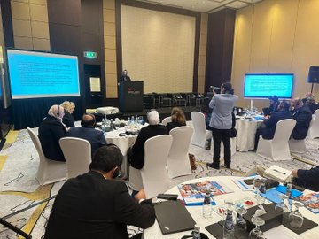 First image displays six men in suits and lanyards seated on a stage with microphones in a conference room featuring beige walls and modern chairs. Second image shows a projector screen with Turkish text about education for Syrian students a male speaker at podium and audience members including women in headscarves seated around tables in a hotel ballroom with Marriott branding. Third image captures a workshop session with diverse attendees in business attire at round tables with laptops documents and water bottles under a chandelier in a spacious room with multiple screens displaying text. Fourth image depicts another meeting with participants seated at tables facing dual screens showing Arabic text a man speaking at podium and photographers capturing the scene in a elegant venue with beige decor and beverages on tables.