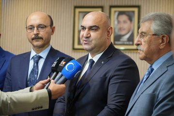 First image shows five men in suits standing in a formal room with striped walls, bookshelves, framed portraits of men, a Turkish flag, and a blue flag with stars, one man holding a red box with a white crescent and star emblem, seated chairs and a table in the background. Second image depicts four men in suits in a room with striped walls and two framed portraits, one bald man handing a small trophy to another man with glasses, a small plant on a table nearby. Third image displays a group of about twelve men in suits seated around low tables in a spacious light-floored room with beige walls, a shuttered window, paintings, plants, a bookshelf, and flags including a red one with white emblem and a blue one with stars. Fourth image features four men in suits standing in a room with striped walls and framed portraits, microphones with TRT and EU logos held toward a bald man speaking, another man holding a microphone.