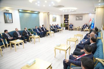 First image shows five men in suits standing in a formal room with striped walls, bookshelves, framed portraits of men, a Turkish flag, and a blue flag with stars, one man holding a red box with a white crescent and star emblem, seated chairs and a table in the background. Second image depicts four men in suits in a room with striped walls and two framed portraits, one bald man handing a small trophy to another man with glasses, a small plant on a table nearby. Third image displays a group of about twelve men in suits seated around low tables in a spacious light-floored room with beige walls, a shuttered window, paintings, plants, a bookshelf, and flags including a red one with white emblem and a blue one with stars. Fourth image features four men in suits standing in a room with striped walls and framed portraits, microphones with TRT and EU logos held toward a bald man speaking, another man holding a microphone.