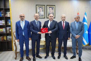 First image shows five men in suits standing in a formal room with striped walls, bookshelves, framed portraits of men, a Turkish flag, and a blue flag with stars, one man holding a red box with a white crescent and star emblem, seated chairs and a table in the background. Second image depicts four men in suits in a room with striped walls and two framed portraits, one bald man handing a small trophy to another man with glasses, a small plant on a table nearby. Third image displays a group of about twelve men in suits seated around low tables in a spacious light-floored room with beige walls, a shuttered window, paintings, plants, a bookshelf, and flags including a red one with white emblem and a blue one with stars. Fourth image features four men in suits standing in a room with striped walls and framed portraits, microphones with TRT and EU logos held toward a bald man speaking, another man holding a microphone.