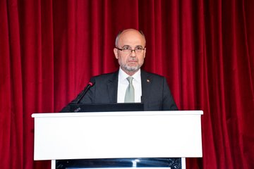 Group photo of diverse participants including men and women in formal attire such as suits jackets and dresses standing together in front of a large screen displaying Yıldızlar Ligi Erasmus+ Merkezi Projeler Kafkas Üniversitesi and Erasmus+ logo with Turkish flag elements and event details for 2025 at an indoor venue. Close-up of a bald man with glasses in a dark suit and green tie speaking at a podium against red curtains. Woman with curly hair in a dark blazer gesturing while speaking at a podium with a laptop screen showing text banners for Yıldızlar Ligi Erasmus+ and Turkish flag in the background against red curtains.