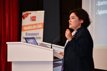 Group photo of diverse participants including men and women in formal attire such as suits jackets and dresses standing together in front of a large screen displaying Yıldızlar Ligi Erasmus+ Merkezi Projeler Kafkas Üniversitesi and Erasmus+ logo with Turkish flag elements and event details for 2025 at an indoor venue. Close-up of a bald man with glasses in a dark suit and green tie speaking at a podium against red curtains. Woman with curly hair in a dark blazer gesturing while speaking at a podium with a laptop screen showing text banners for Yıldızlar Ligi Erasmus+ and Turkish flag in the background against red curtains.