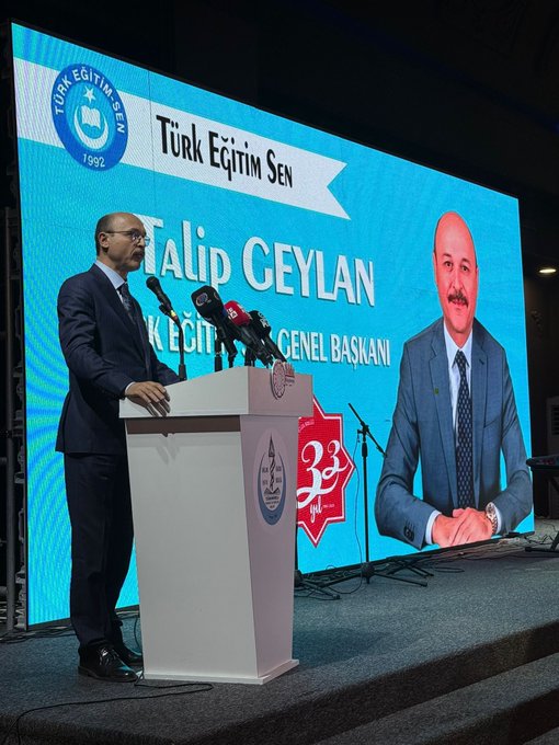 First image shows a man in a dark suit standing at a white podium with microphones on a stage, speaking, with a large blue-lit screen behind displaying Turkish Education Union banner, his name Talip Geylan as general president, and a large photo of him smiling. Second image depicts rows of men in suits seated in an ornate hall with chandeliers and draped ceilings, some holding books or bags, attending a formal gathering. Third image captures a diverse group of men women and children in formal attire seated in a grand venue with blue decorations and flags including Turkmen flag, some holding small flags, in an audience setting. Fourth image shows a large crowd of people standing and seated in a decorated hall with stage screens displaying event graphics, blue lighting, and ornate ceiling, gathered for a formal assembly.