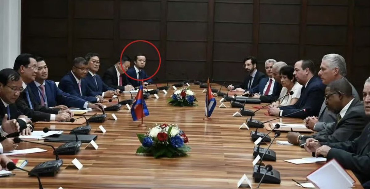 会议室里铺着白布的长木桌，周围是穿着正式西装的男女坐在椅子上，有些人还戴着名牌。中心以红白相间的花朵为中心，桌上挂着包括柬埔寨在内的多个国旗。麦克风和文件散落一地。红色圆圈突出了一名身穿深色西装的亚洲男子坐在人群中，所有人都在讨论。