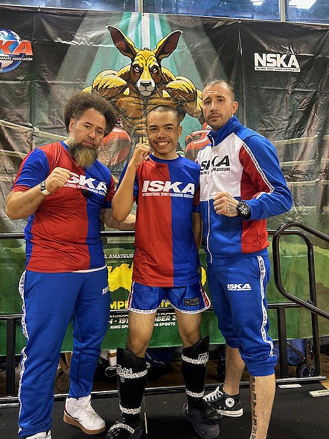 First image shows a young male athlete with short hair smiling confidently while holding a large black and gold championship belt over his shoulder with one hand raised in a fist pump. He wears a blue and white jacket with red accents and stands against a blue background with shattered glass effects around his fists. Second image depicts three men on a stage with an ISKA banner featuring a kangaroo mascot. The central figure is the young athlete in red and blue shorts and a matching top posing with fists up. Flanking him are two older men one with a beard in blue pants and jacket and the other in a blue tracksuit both wearing ISKA branded clothing. The setting includes a podium and green railing.