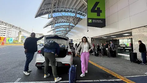 People stand near a white car with its trunk open at an airport terminal. A man in dark clothing points toward the trunk while another person in pink pants and a white top stands nearby holding a suitcase. Luggage bags are visible on the ground. A large green sign with the number 4 is attached to a modern canopy structure overhead. The scene includes other travelers and vehicles in the background under a clear sky.