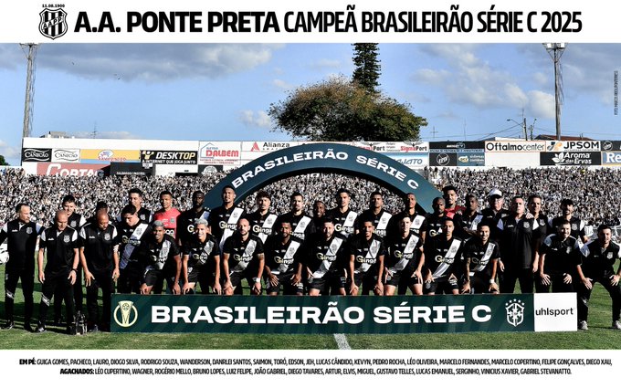 Group of male soccer players in black and white uniforms standing together on a green field in a stadium with stands in background under bright lights and blue sky, central banner arch reading A.A. Ponte Preta Campeao Brasileiro Serie C 2025, lower banner with Brasileirao Serie C and sponsor Uninorte, team members including names like Douglas Baggio, Anderson Leite, and others listed below.