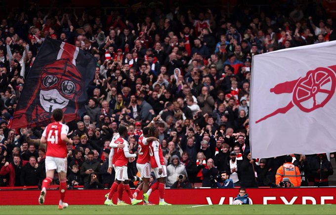 First image shows a man with dark hair and beard wearing a black puffer jacket with collar up, standing on a football field sideline with arms crossed, looking serious amid a crowd of spectators in the background wearing red and white Arsenal kits. Second image depicts a group of Arsenal players in red and white uniforms celebrating on the green pitch, with fans in the stands holding a large banner featuring a masked face design in black and red, and the Arsenal logo visible on a white flag, surrounded by cheering supporters and stadium seating.