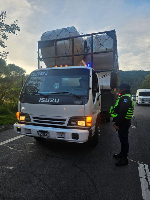 Primera imagen muestra un camion blanco con contenedores de carga en una carretera rural bajo cielo azul con arboles al fondo dos policias con cascos y chalecos verdes estan cerca del camion uno inspecciona el area y otro esta al lado del vehiculo la carretera tiene lineas blancas y un pequeno monticulo de tierra al lado. Segunda imagen muestra un camion Isuzu blanco con luces azules activas y dos grandes contenedores de carga envueltos en malla un policia con chaleco reflectante verde esta parado al lado del camion en una carretera asfaltada con vegetacion y otro vehiculo blanco visible al fondo el camion tiene placa visible y esta estacionado en el borde de la via.