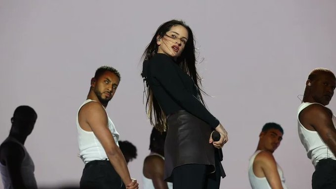 Rosalía stands center stage in a black long-sleeve top and black leather skirt, holding a microphone, with long dark hair and a focused expression. Several male dancers in white tank tops and dark pants surround her, posing dynamically with arms extended. The background is a solid purple with shadowy silhouettes.
