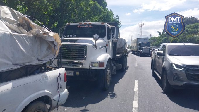 A white dump truck with license plate C-IRON stopped on the right side of a two-lane road, covered by a blue tarp, positioned near the edge with its rear extending into the lane. The truck has a front grille and side mirrors visible, with mechanical issues implied by the context. Surrounding vehicles include a gray pickup truck and a semi-truck in the background, along with trees and a blue sign displaying PMT logo on the roadside under a partly cloudy sky.