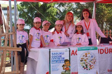 First image shows group of young girls wearing pink shirts and hats with school logo standing at outdoor booth under red canopy with pine trees in background, table covered in white cloth displaying nutrition banner titled SAGLIKLI ACILIK SAGLIKLI GELECEK featuring cartoon child and balanced food plate illustration including vegetables fruits grains proteins and dairy. Second image depicts girl in white lab coat and pink hat using stethoscope on boys chest while he sits in red and gray shirt, other children in similar costumes around them in sunny outdoor setting with chairs. Third image captures children in red shirts holding books near red tent labeled TRABZON IL SAGLIK MUDURLUGU with banner SAGLIKLI COCUK SAGLIKLI GELECEK above tooth symbol and heart, adults including man in yellow shirt and woman in pink hat at table with posters on volunteering and health awareness, parked van nearby. Fourth image features two girls in white lab coats holding certificates with health project logo and red ribbon, smiling boy in white hat and shirt next to them, adult man in suit and glasses and woman in red scrubs standing behind group against red wall.