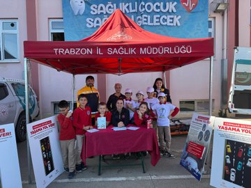 First image shows group of young girls wearing pink shirts and hats with school logo standing at outdoor booth under red canopy with pine trees in background, table covered in white cloth displaying nutrition banner titled SAGLIKLI ACILIK SAGLIKLI GELECEK featuring cartoon child and balanced food plate illustration including vegetables fruits grains proteins and dairy. Second image depicts girl in white lab coat and pink hat using stethoscope on boys chest while he sits in red and gray shirt, other children in similar costumes around them in sunny outdoor setting with chairs. Third image captures children in red shirts holding books near red tent labeled TRABZON IL SAGLIK MUDURLUGU with banner SAGLIKLI COCUK SAGLIKLI GELECEK above tooth symbol and heart, adults including man in yellow shirt and woman in pink hat at table with posters on volunteering and health awareness, parked van nearby. Fourth image features two girls in white lab coats holding certificates with health project logo and red ribbon, smiling boy in white hat and shirt next to them, adult man in suit and glasses and woman in red scrubs standing behind group against red wall.
