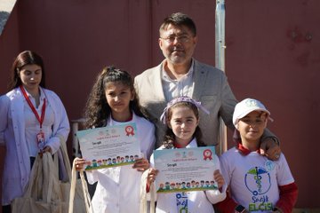 First image shows group of young girls wearing pink shirts and hats with school logo standing at outdoor booth under red canopy with pine trees in background, table covered in white cloth displaying nutrition banner titled SAGLIKLI ACILIK SAGLIKLI GELECEK featuring cartoon child and balanced food plate illustration including vegetables fruits grains proteins and dairy. Second image depicts girl in white lab coat and pink hat using stethoscope on boys chest while he sits in red and gray shirt, other children in similar costumes around them in sunny outdoor setting with chairs. Third image captures children in red shirts holding books near red tent labeled TRABZON IL SAGLIK MUDURLUGU with banner SAGLIKLI COCUK SAGLIKLI GELECEK above tooth symbol and heart, adults including man in yellow shirt and woman in pink hat at table with posters on volunteering and health awareness, parked van nearby. Fourth image features two girls in white lab coats holding certificates with health project logo and red ribbon, smiling boy in white hat and shirt next to them, adult man in suit and glasses and woman in red scrubs standing behind group against red wall.