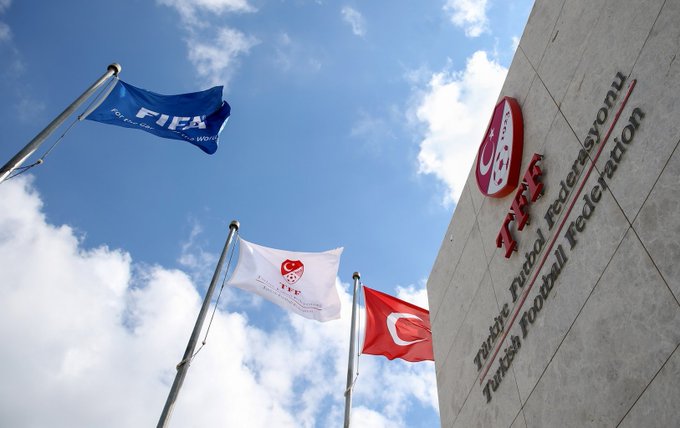 Building facade displays Turkish Football Federation text. Flags of FIFA in blue UEFA in red and white Turkish in red and white wave on poles against clear blue sky with scattered clouds.
