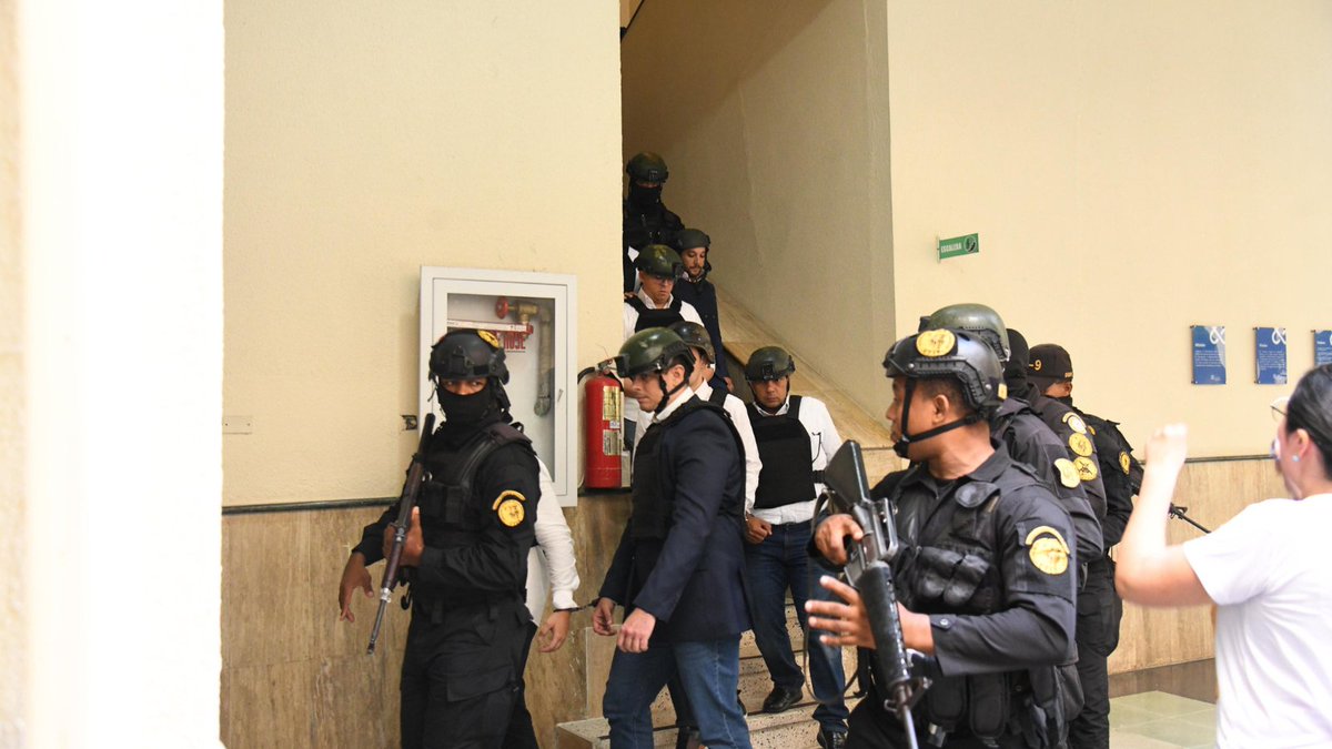 Group of police officers in black tactical uniforms, helmets, and masks, some carrying rifles, positioned near stairs and a doorway in a building interior with beige walls, a fire extinguisher cabinet, and green exit sign. One officer in suit stands among them, and a woman in white shirt is visible nearby.