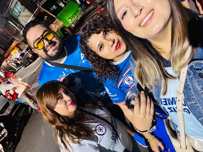 Group of four people standing on a street at night near a green bus and vehicles. Two men wear blue jerseys with star emblem and sunglasses one holds a can. Two women in casual tops one gray with UNAM patch and jeans smiling at camera. Background includes streetlights and urban setting.