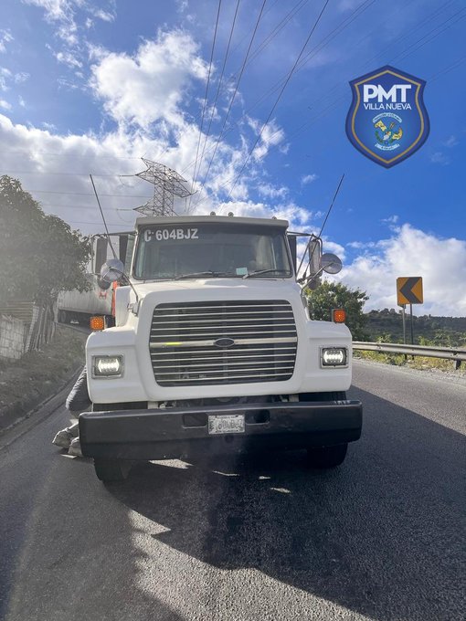 White truck with license plate C 5048 BZ stopped on a roadside uphill under partly cloudy skies, surrounded by greenery and a yellow road sign, with a police emblem visible in the corner.
