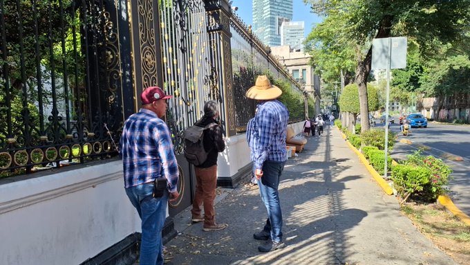 First image displays three men standing near a tall black wrought-iron fence with gold accents surrounding a modern high-rise building partially visible in the background under a clear sky. One man wears a red cap and plaid shirt another a wide-brimmed straw hat and blue plaid shirt the third a black backpack and dark clothing. They appear to be conversing or waiting on a shaded sidewalk lined with green bushes and trees with a street and yellow curb visible. Second image shows a larger group of about 15-20 people including men and women in casual outfits like hats backpacks and light jackets gathered along a similar ornate fence outside a classical-style building with arched windows and topiaries. Some carry bags others stand with hands in pockets on a sunny street with cars parked and greenery bordering the area.