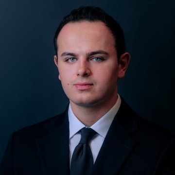 A professional headshot portrait of a young man with short dark hair, light skin, green eyes, wearing a dark suit, white dress shirt, and dark tie, posed against a plain dark blue-gray background.