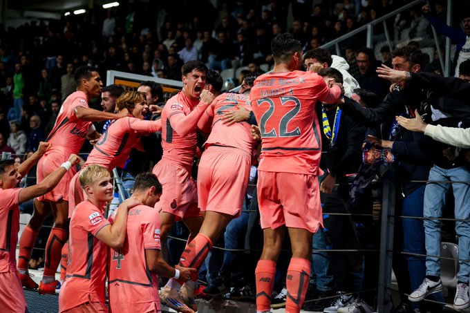 Group of male soccer players in pink and black uniforms with number 22 on one jersey, embracing and gesturing excitedly towards a stadium crowd. Spectators in green and blue clothing fill the stands behind a barrier. The scene shows celebration after a goal in an indoor arena with bright lighting.