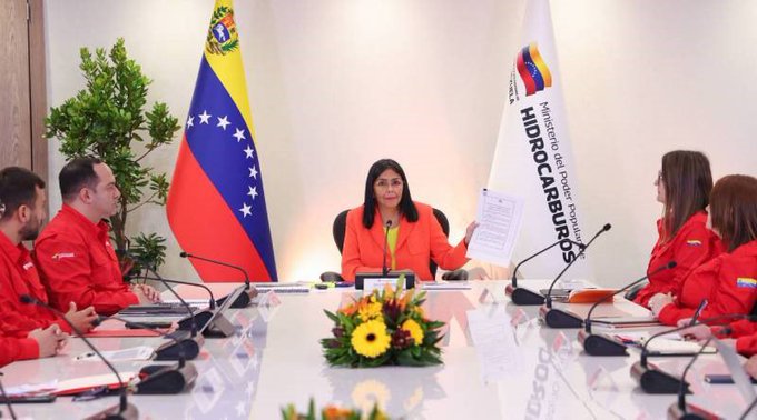 A group of officials in red attire sits around a white conference table in a formal meeting room, with a central woman in orange holding documents, flanked by men and women, Venezuelan and Colombian flags on stands behind, potted plant and yellow flower arrangement on the table, microphones and papers present.