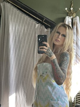 First image shows a blonde woman with heavy makeup and long hair holding a smartphone taking a selfie in a bathroom with green walls, white shower curtain, and brass fixture, wearing a light blue patterned towel wrapped around her body, visible tattoos on her arms. Second image depicts the same woman with ponytail hairstyle holding a smartphone for a selfie in a tiled bathroom with toilet and gray chair, dressed in a gray tank top and black elephant print shorts, tattoos on arms visible. Third image features the woman lying on a bed with gray pillows, smiling with makeup, wearing a chain necklace, tattoos on arms, next to a cartoon orange chipmunk character in a brown hat holding a nut, edited overlay on the image.