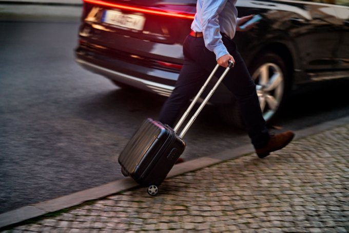A man in a light blue shirt and dark pants walks on a paved urban street pulling a black rolling suitcase with wheels. Behind him is a black luxury car with red taillights illuminated and a license plate visible on the rear. The scene appears to be at dusk or night with street lighting and cobblestone elements nearby.