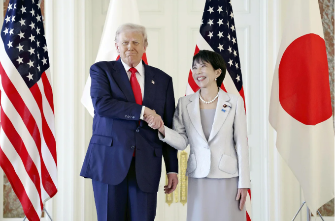 Two leaders stand in a formal room with American and Japanese flags on either side. The man on the left wears a dark suit with a red tie and has white hair, extending his right hand for a shake. The woman on the right wears a light gray suit and pearl necklace, grasping his hand with her right hand while smiling. Both appear engaged in a diplomatic greeting at the State Guest House.