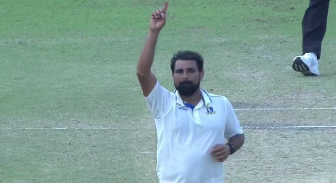 A man with a beard wearing a white collared shirt with blue accents and wristwatch stands on a green grass field raising his right arm with index finger pointed upward in celebration while another persons lower body in black pants and white shoes is visible in the background.