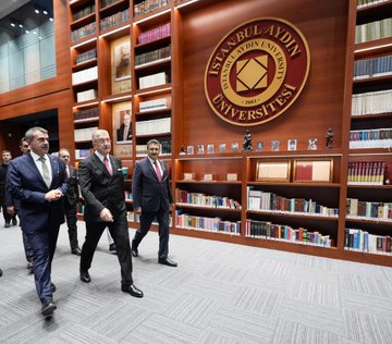 First image shows a modern library interior at Istanbul Aydin University with extensive wooden bookshelves filled with books and a large circular emblem featuring the university name in Turkish on a wall. Several suited men including those in formal attire walk in the foreground near display cases with awards and busts. Second image depicts a large curved auditorium with tiered red seating filled with an audience wearing academic caps and gowns listening to a speaker at a podium on stage under bright lighting and acoustic panels. Third image captures a group of graduates in maroon academic robes and caps standing on a stage with a projector screen in background posing formally after ceremony with flowers nearby. Fourth image shows a diverse group of people including officials and athletes in red tracksuits holding gold medals and trophies on a stage with a banner reading 2025 Istanbul Aydin University Sports Awards in Turkish surrounded by Turkish flags.