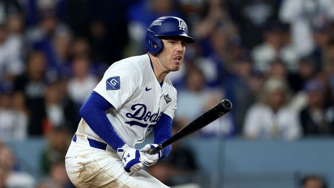A male baseball player in a white Los Angeles Dodgers uniform with blue accents and protective helmet swings a bat during a game with a crowd in blue attire visible in the background stadium seating.