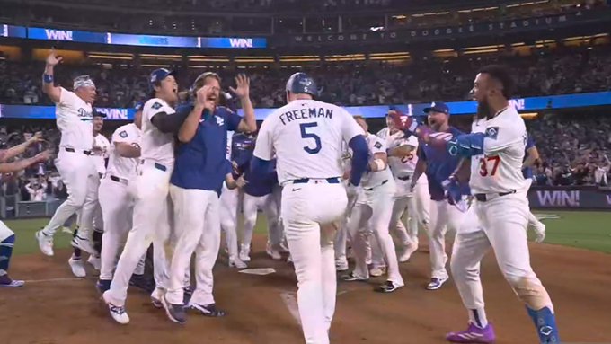 Group of Los Angeles Dodgers players in white and blue uniforms celebrating a victory on the baseball field at night under stadium lights, with arms raised and some jumping, scoreboard showing WIN in the background, player with FREEMAN on jersey number 5 visible from behind.