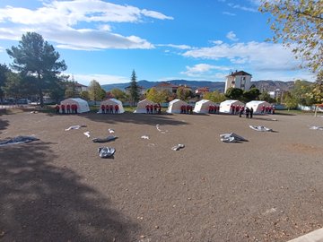 First image shows large group of people mostly in red jackets and white helmets posing together outdoors on gravel ground with some kneeling, behind them several white tents labeled AFAD, trees and a beige building with tower in background under partly cloudy blue sky. Second image displays multiple white AFAD tents arranged in open dirt area with scattered fabric pieces on ground, groups of people in red attire standing near tents, trees and mountains in distant background under clear blue sky. Third image features another group of individuals in red uniforms and helmets sitting and standing on green grass near white AFAD tents, autumn trees visible in background under cloudy sky.