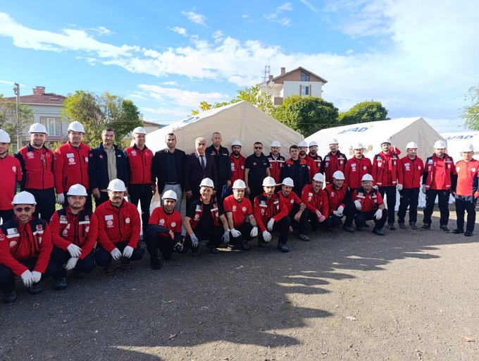 First image shows large group of people mostly in red jackets and white helmets posing together outdoors on gravel ground with some kneeling, behind them several white tents labeled AFAD, trees and a beige building with tower in background under partly cloudy blue sky. Second image displays multiple white AFAD tents arranged in open dirt area with scattered fabric pieces on ground, groups of people in red attire standing near tents, trees and mountains in distant background under clear blue sky. Third image features another group of individuals in red uniforms and helmets sitting and standing on green grass near white AFAD tents, autumn trees visible in background under cloudy sky.