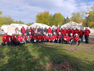 First image shows large group of people mostly in red jackets and white helmets posing together outdoors on gravel ground with some kneeling, behind them several white tents labeled AFAD, trees and a beige building with tower in background under partly cloudy blue sky. Second image displays multiple white AFAD tents arranged in open dirt area with scattered fabric pieces on ground, groups of people in red attire standing near tents, trees and mountains in distant background under clear blue sky. Third image features another group of individuals in red uniforms and helmets sitting and standing on green grass near white AFAD tents, autumn trees visible in background under cloudy sky.