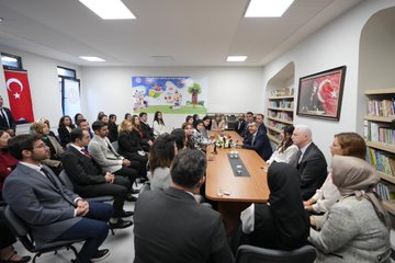First image shows a man in suit kneeling and hugging two young girls in school uniforms with backpacks, one wearing a red hooded cape and headband with crescent moon, smiling together outdoors with other suited men in background. Second image depicts a man in suit speaking at a podium on a stage with blue backdrop featuring Turkish flags, Atatürk portrait, and text Gümüşdere İlkokulu ve Ortaokulu Açılış Töreni, audience seated in front. Third image captures a large group of adults and children in formal attire cutting a ribbon under balloon arch with Gümüşdere Okulu sign at building entrance. Fourth image shows diverse group of suited men and women, some with headscarves, seated around a wooden table in a library with bookshelves, Turkish flags, childrens drawings on wall, and vase of flowers.