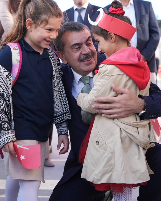 First image shows a man in suit kneeling and hugging two young girls in school uniforms with backpacks, one wearing a red hooded cape and headband with crescent moon, smiling together outdoors with other suited men in background. Second image depicts a man in suit speaking at a podium on a stage with blue backdrop featuring Turkish flags, Atatürk portrait, and text Gümüşdere İlkokulu ve Ortaokulu Açılış Töreni, audience seated in front. Third image captures a large group of adults and children in formal attire cutting a ribbon under balloon arch with Gümüşdere Okulu sign at building entrance. Fourth image shows diverse group of suited men and women, some with headscarves, seated around a wooden table in a library with bookshelves, Turkish flags, childrens drawings on wall, and vase of flowers.