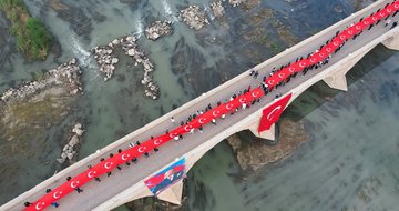 Aerial view of a long red carpet laid across an ancient stone bridge over a greenish river with numerous people in dark clothing walking along it holding a massive Turkish flag draped over the side and smaller flags waving nearby surrounded by rocky banks and sparse vegetation. Another aerial shot shows a red carpet extending through a historic town square with terracotta-roofed buildings cars parked along streets and groups of people walking amid trees and open spaces. Overhead image captures a crowd of people on a bridge over reflective water with mountains in background holding large red Turkish flags including one featuring a portrait of Mustafa Kemal Ataturk with a minaret visible in distance. Final aerial perspective displays participants on a bridge spanning a blue-green river with red carpet and Turkish flags billowing in wind against stone structure and waterside greenery.