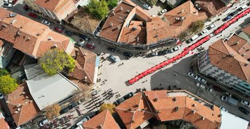 Aerial view of a long red carpet laid across an ancient stone bridge over a greenish river with numerous people in dark clothing walking along it holding a massive Turkish flag draped over the side and smaller flags waving nearby surrounded by rocky banks and sparse vegetation. Another aerial shot shows a red carpet extending through a historic town square with terracotta-roofed buildings cars parked along streets and groups of people walking amid trees and open spaces. Overhead image captures a crowd of people on a bridge over reflective water with mountains in background holding large red Turkish flags including one featuring a portrait of Mustafa Kemal Ataturk with a minaret visible in distance. Final aerial perspective displays participants on a bridge spanning a blue-green river with red carpet and Turkish flags billowing in wind against stone structure and waterside greenery.