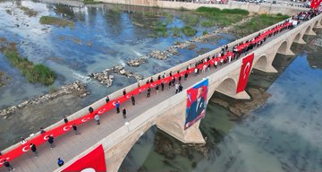 Aerial view of a long red carpet laid across an ancient stone bridge over a greenish river with numerous people in dark clothing walking along it holding a massive Turkish flag draped over the side and smaller flags waving nearby surrounded by rocky banks and sparse vegetation. Another aerial shot shows a red carpet extending through a historic town square with terracotta-roofed buildings cars parked along streets and groups of people walking amid trees and open spaces. Overhead image captures a crowd of people on a bridge over reflective water with mountains in background holding large red Turkish flags including one featuring a portrait of Mustafa Kemal Ataturk with a minaret visible in distance. Final aerial perspective displays participants on a bridge spanning a blue-green river with red carpet and Turkish flags billowing in wind against stone structure and waterside greenery.